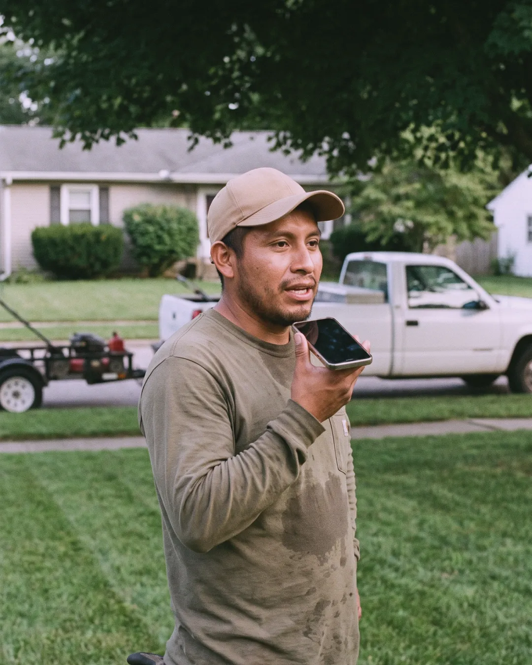 A landscaper in a cap records a job into his phone in front of his truck